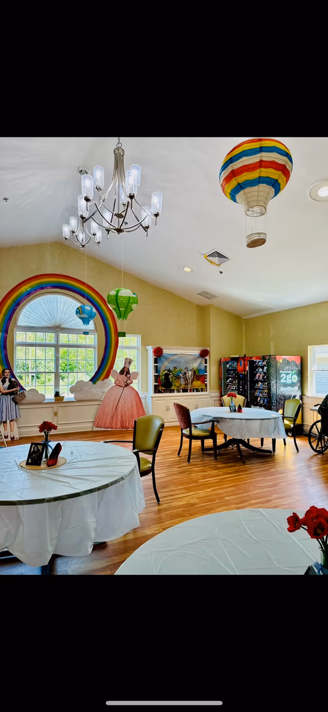 Bright common dining/activity room with round tables covered in white cloths, a rainbow wall mural, hanging hot-air-balloon decorations, and vending machines along the back wall.