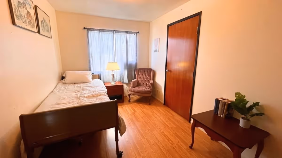 Small furnished bedroom with a single bed, nightstand and lamp by a window, an upholstered armchair, wooden door, and a side table with books and a plant.