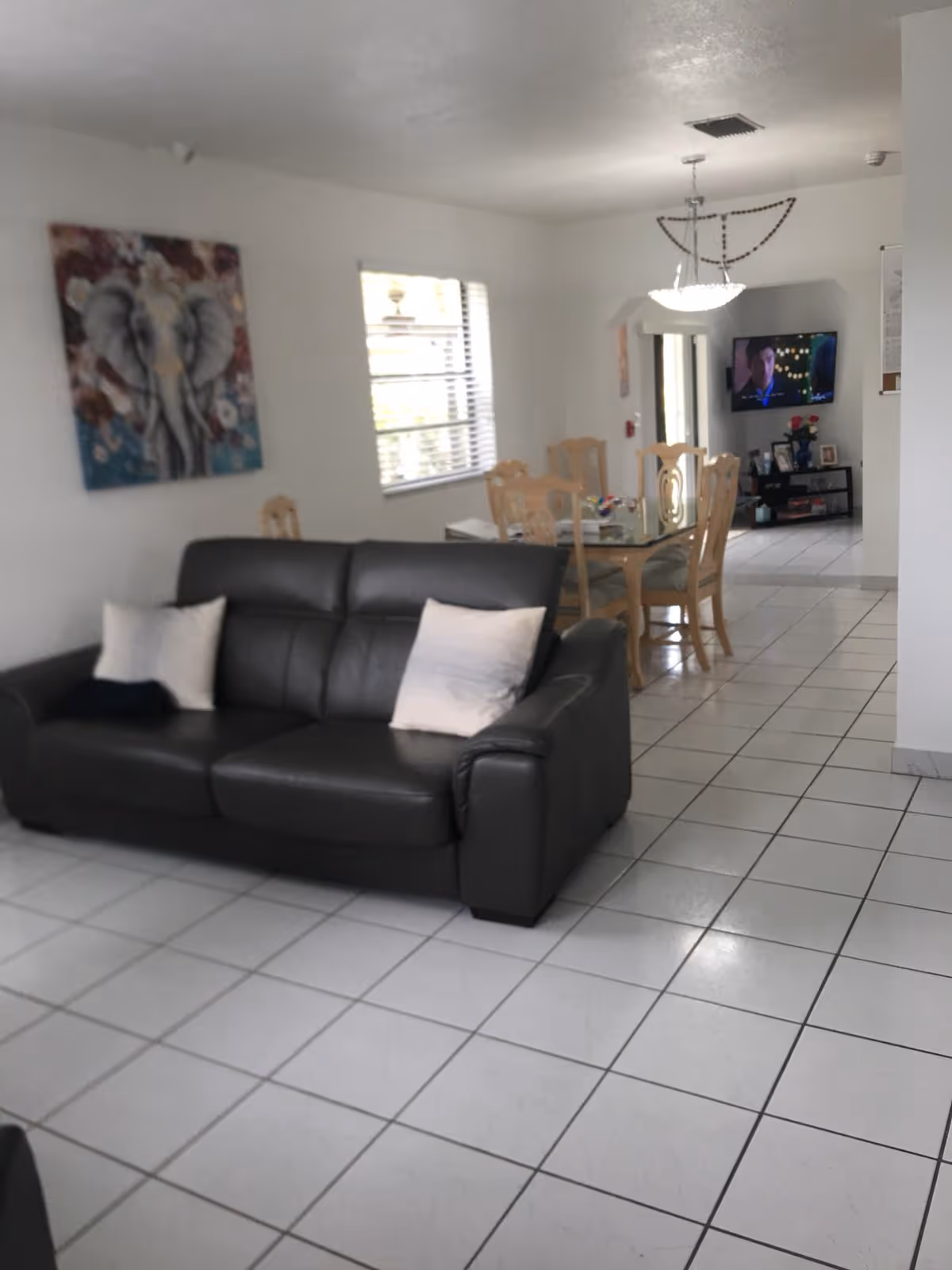 Open-plan living and dining area with a dark leather sofa, tiled floor, dining table and wall-mounted TV.
