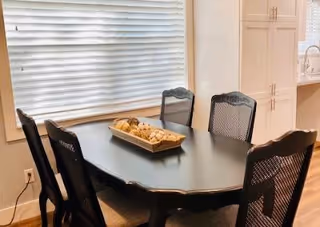 A dining area with a black oval dining table surrounded by six black chairs with mesh backs. On the table is a rectangular tray filled with decorative items. The room has a large window with white blinds and white cabinetry in the background.