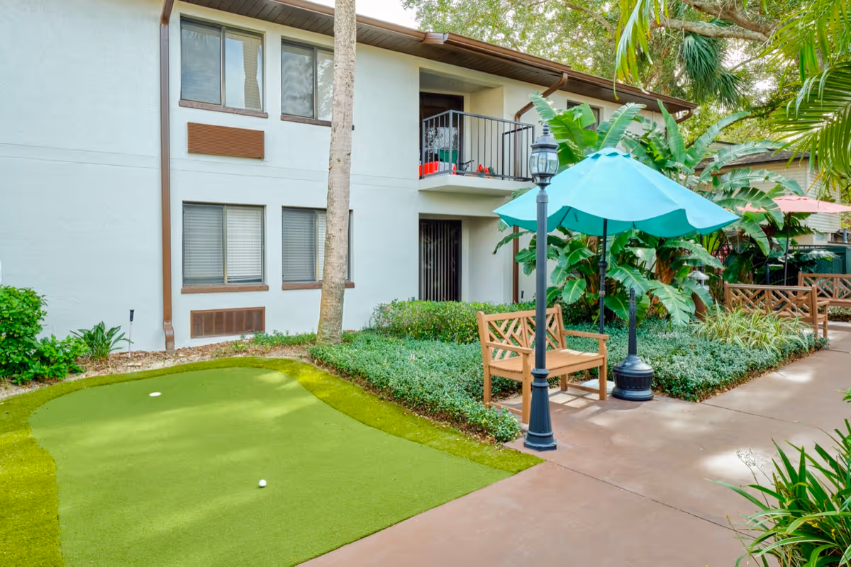 Outdoor area of a senior living facility with a small artificial putting green, a wooden bench under a teal umbrella, a black lamp post, and lush green plants surrounding a concrete walkway. The building exterior is visible with windows and a balcony on the second floor.