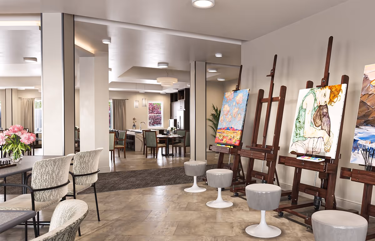 Interior view of a senior living facility art room with several wooden easels holding colorful paintings. There are round stools in front of the easels and a table with chairs and a vase of flowers. In the background, a dining area with tables and chairs is visible, along with a kitchen area.