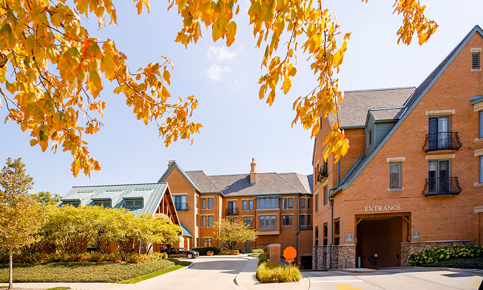 Exterior view of a senior living facility building with brick walls and multiple windows, surrounded by trees with autumn-colored leaves under a clear blue sky. The building has an entrance archway and a driveway leading up to it.