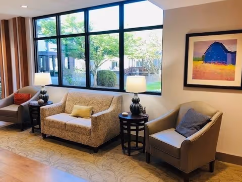 A cozy seating area with a beige loveseat and two armchairs, each with a cushion. Two side tables with lamps are placed beside the loveseat and one armchair. Large windows behind the seating area show greenery outside. A colorful framed painting of a barn hangs on the wall.