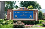 A brick and stone sign at the entrance of Masonic Care Community of New York, surrounded by green shrubs, flowers, and trees in the background.