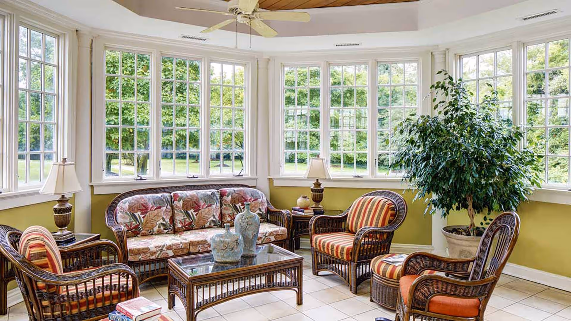 A bright sunroom with large windows on three sides showing green trees outside. The room has wicker furniture including a floral-patterned sofa, three armchairs with striped cushions, a glass-top coffee table with two decorative vases, two side tables with lamps, and a large potted plant in the corner. The ceiling has a white fan and the walls are painted light green with white trim.