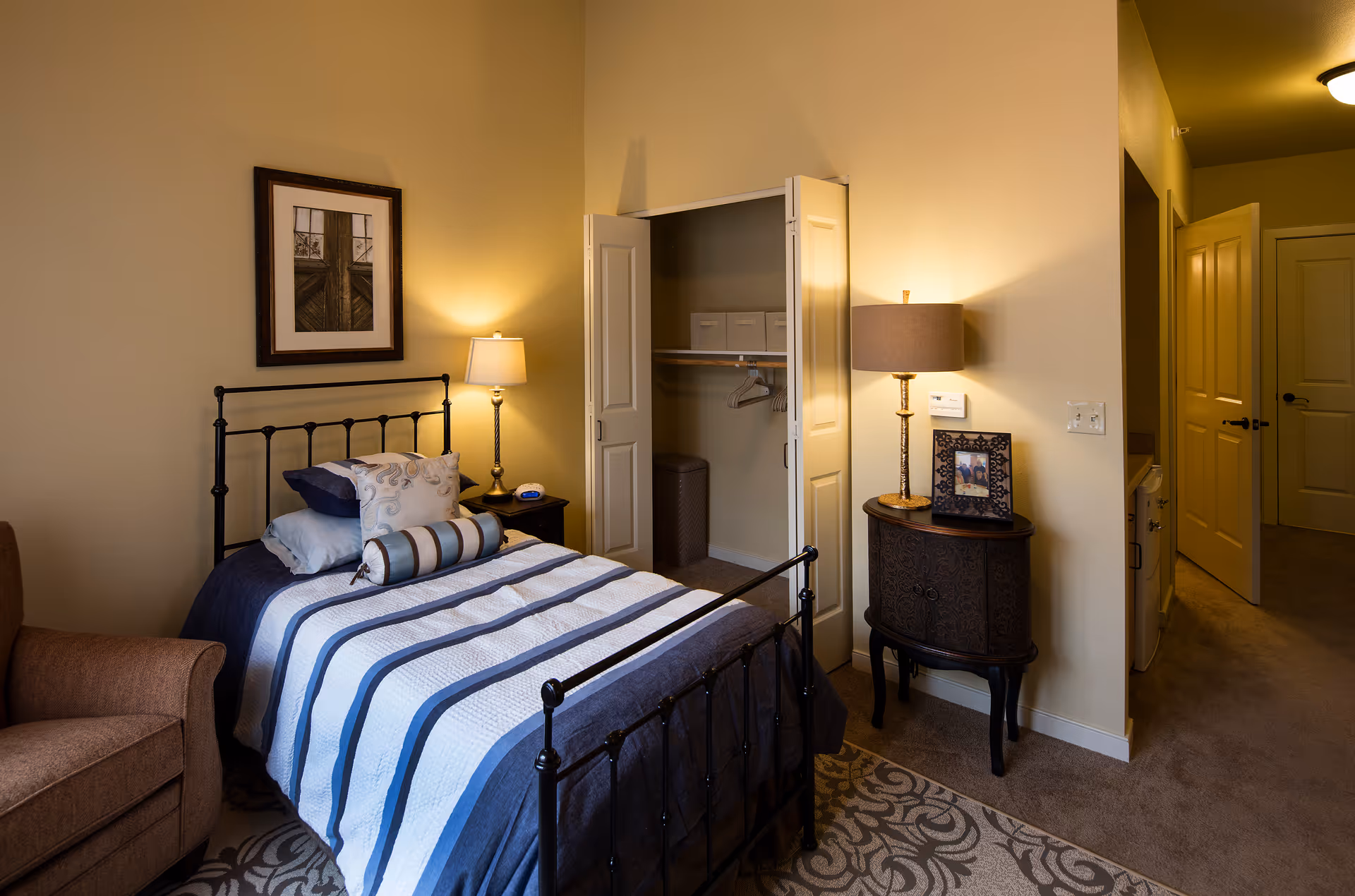 A cozy bedroom in a senior living facility featuring a bed with a striped blue and white quilt, decorative pillows, a black metal headboard, a nightstand with a lamp and alarm clock, an open closet with hangers and storage boxes, a small ornate cabinet with a lamp and framed photo, a brown armchair, and beige walls and carpet.