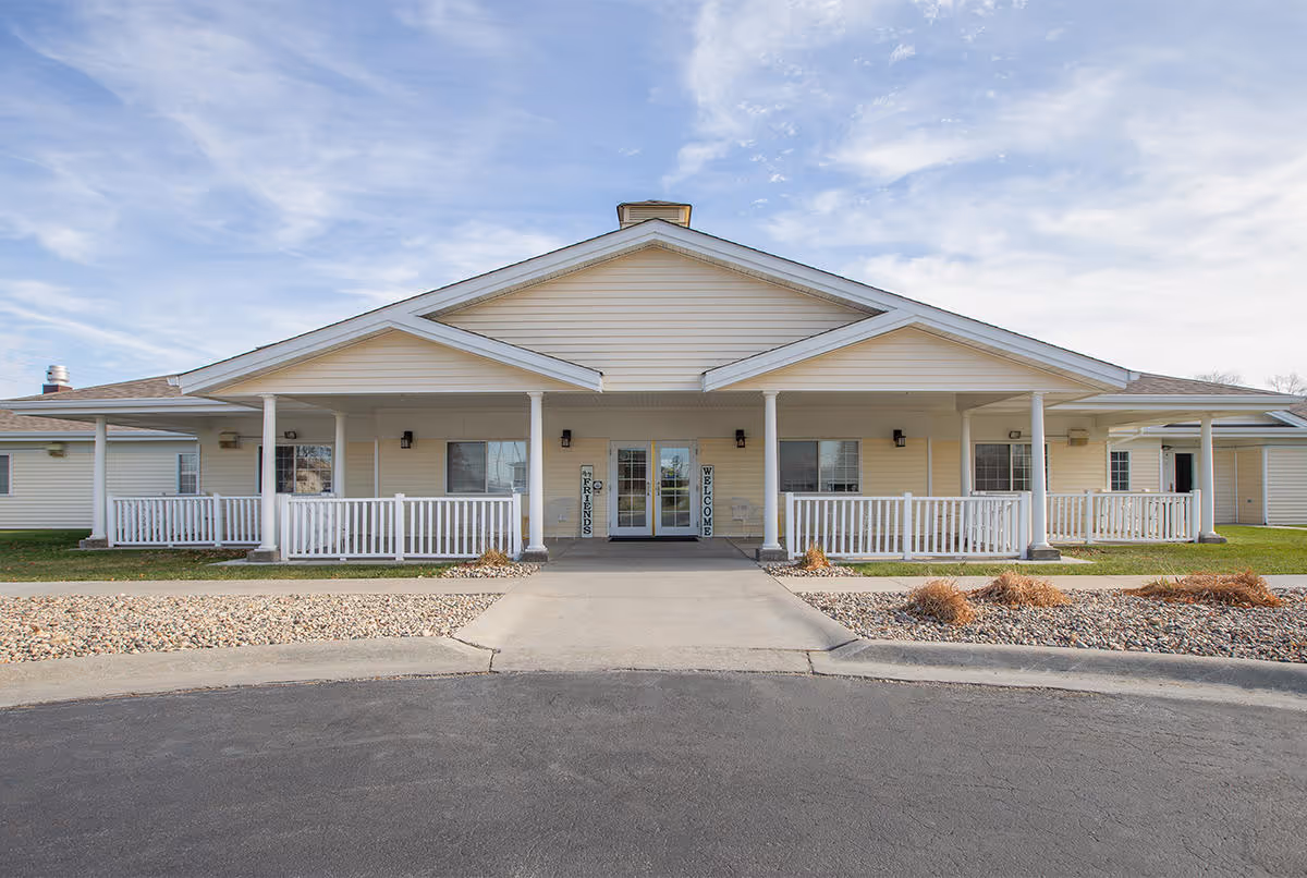 Front exterior view of a single-story senior living facility building with a covered entrance, white railings, and a welcoming sign on either side of the double glass doors. The sky is partly cloudy.