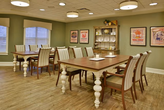 A dining room in a senior living facility with two wooden tables, each surrounded by eight upholstered chairs with striped fabric. The room has green walls, three framed vintage-style posters, a wooden hutch with plates and decorative items, and large windows with blinds. The floor is wood, and the ceiling has recessed lighting and round light fixtures.