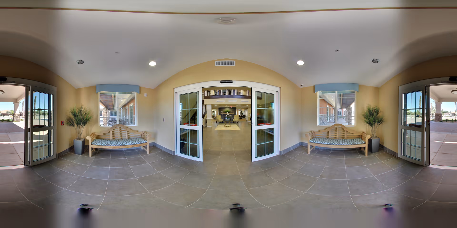 Entry lobby with automatic sliding glass doors, two wooden benches with cushions and potted plants, leading into a reception area.