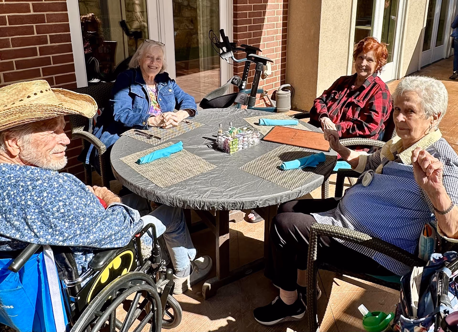 Four elderly people sitting around a round outdoor table with placemats and blue napkins, enjoying a sunny day. One man in a wheelchair wearing a cowboy hat and a blue patterned shirt, and three women, one in a denim jacket, one in a red plaid shirt, and one in a blue and white striped shirt. The setting appears to be a patio area outside a building with brick and beige walls.