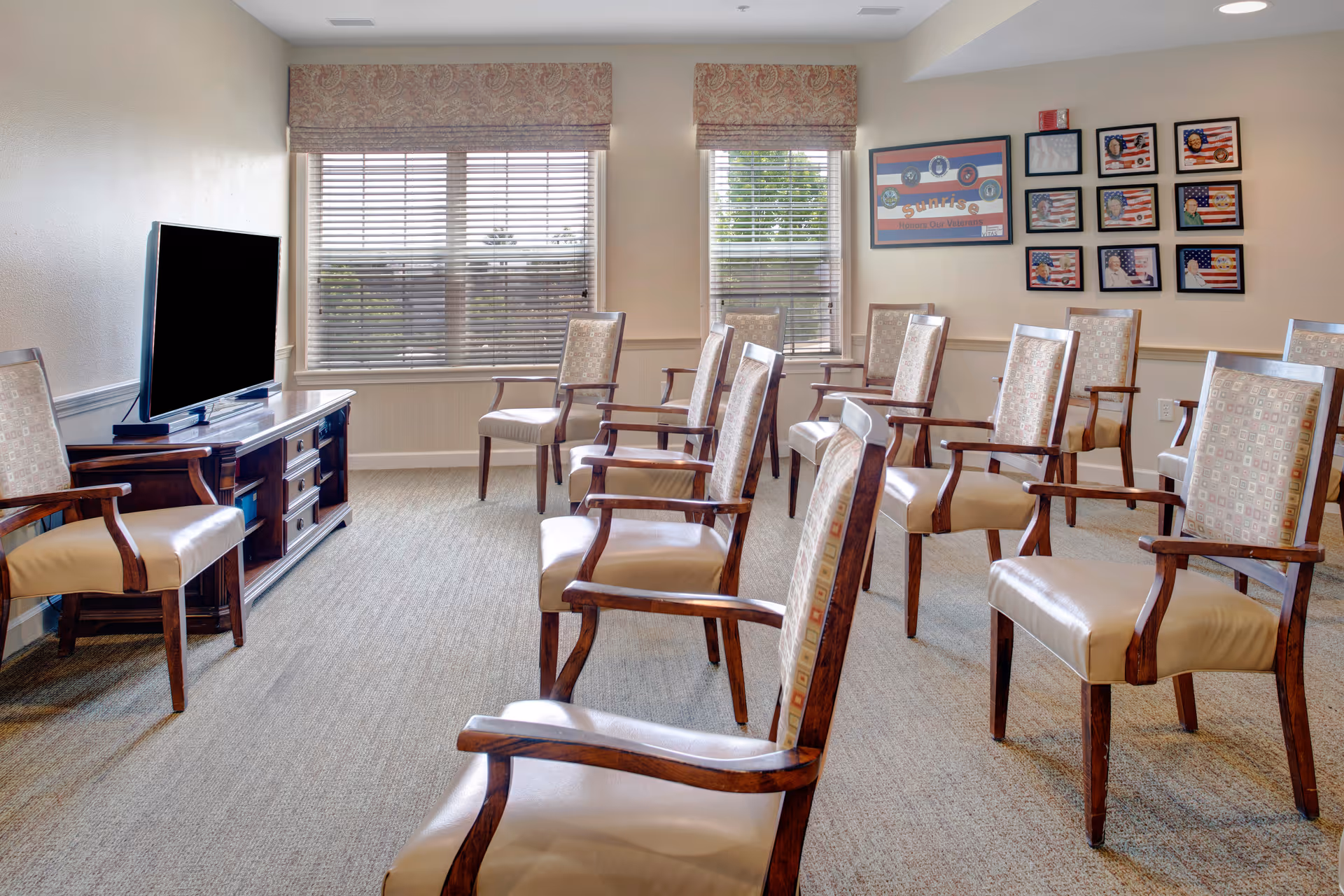 A senior living common room with rows of upholstered wooden chairs arranged facing a television and large windows.