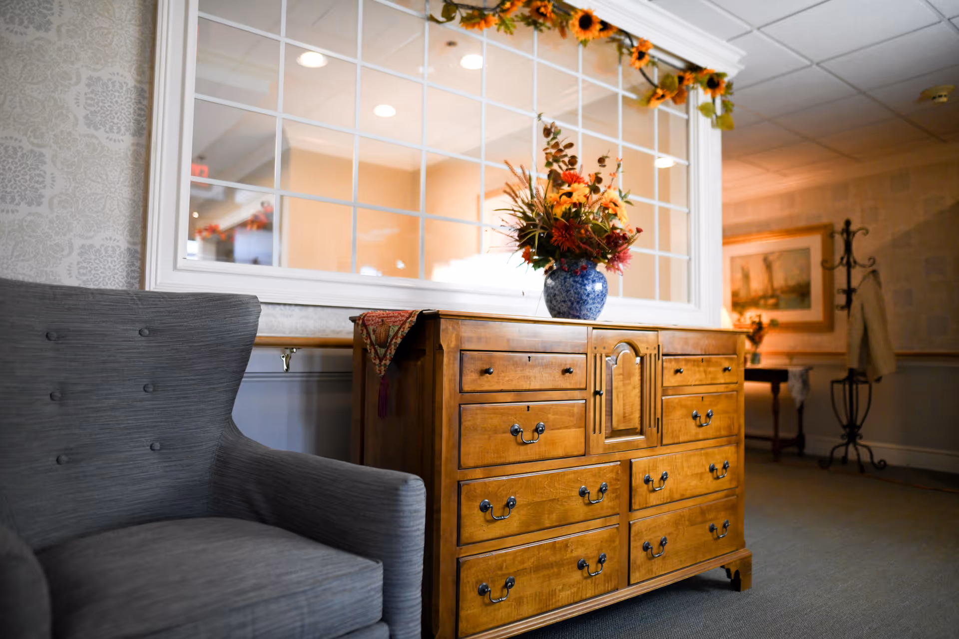 A cozy interior corner featuring a gray upholstered armchair next to a wooden dresser with multiple drawers. On top of the dresser is a blue and white vase filled with a colorful autumn-themed flower arrangement. Behind the dresser is a large window with multiple panes, decorated with a garland of sunflowers. The background includes a coat rack with a beige coat and a framed painting on the wall.