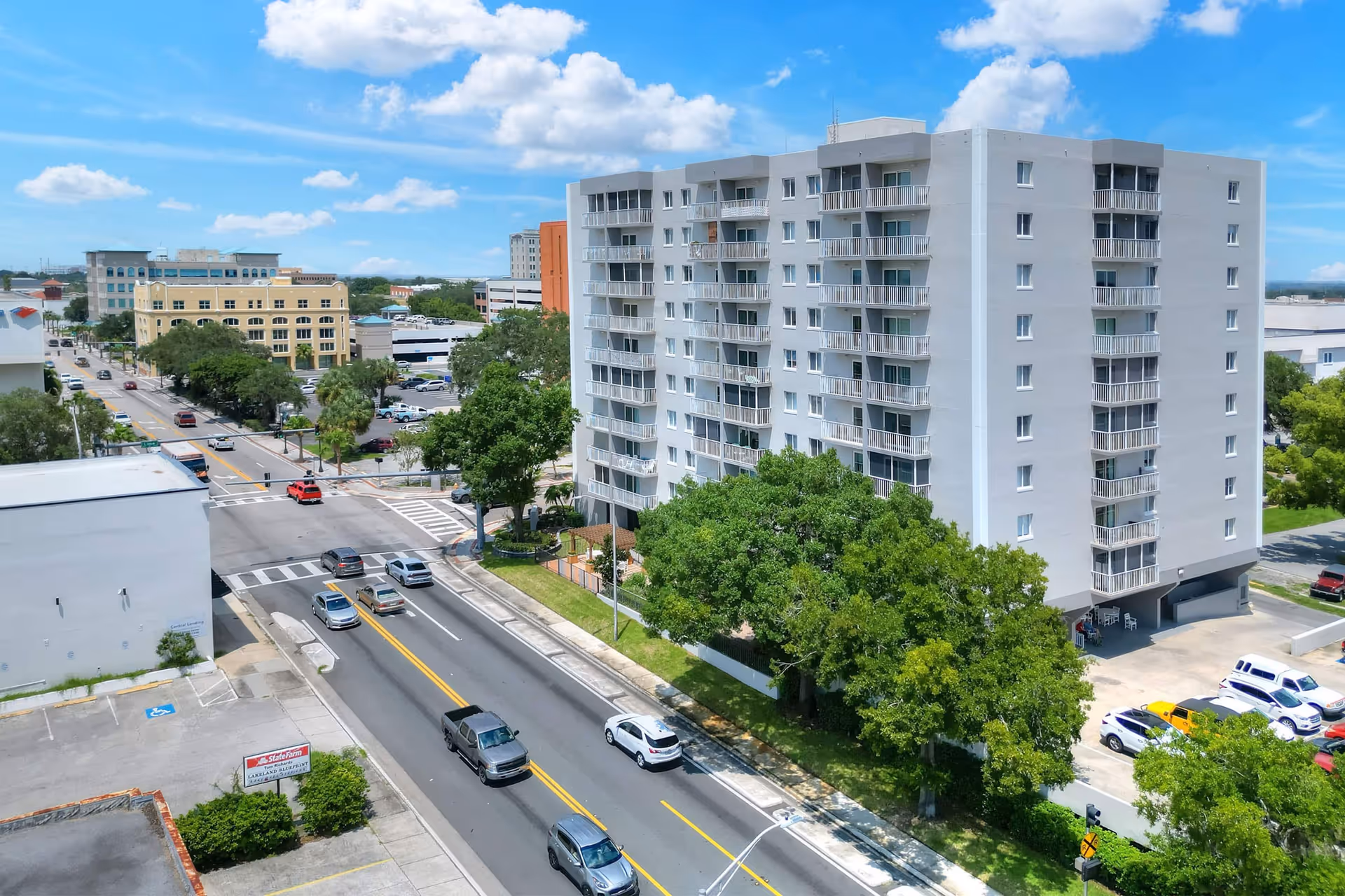 A multi-story white residential building with balconies on each floor, surrounded by trees and adjacent to a busy street with cars. The sky is blue with scattered clouds, and other buildings are visible in the background.