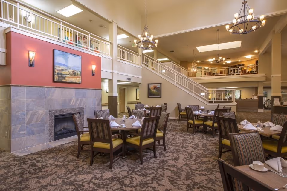 Spacious dining area in a senior living facility with multiple tables set with white napkins and cups. The room features a fireplace with a painting above it, patterned carpet, and a staircase leading to an upper level with railings. Several chandeliers hang from the ceiling providing warm lighting.