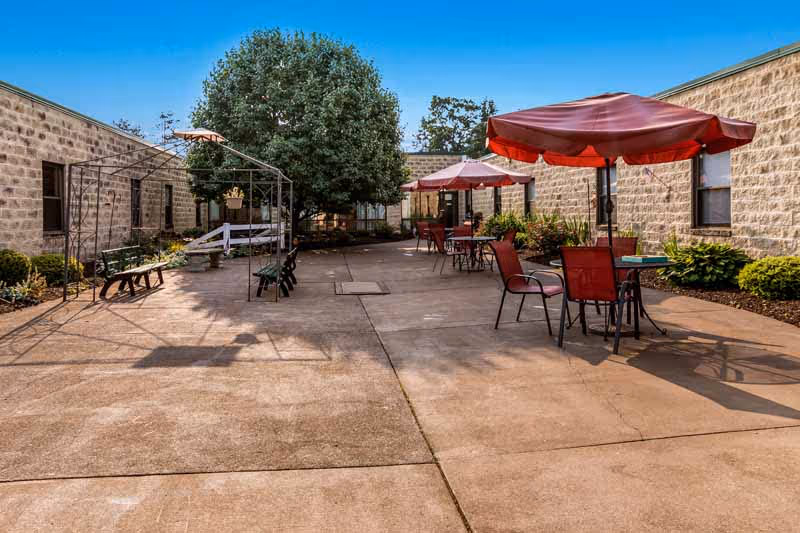 Outdoor courtyard with patio tables and red umbrellas between low stone buildings and a central tree.