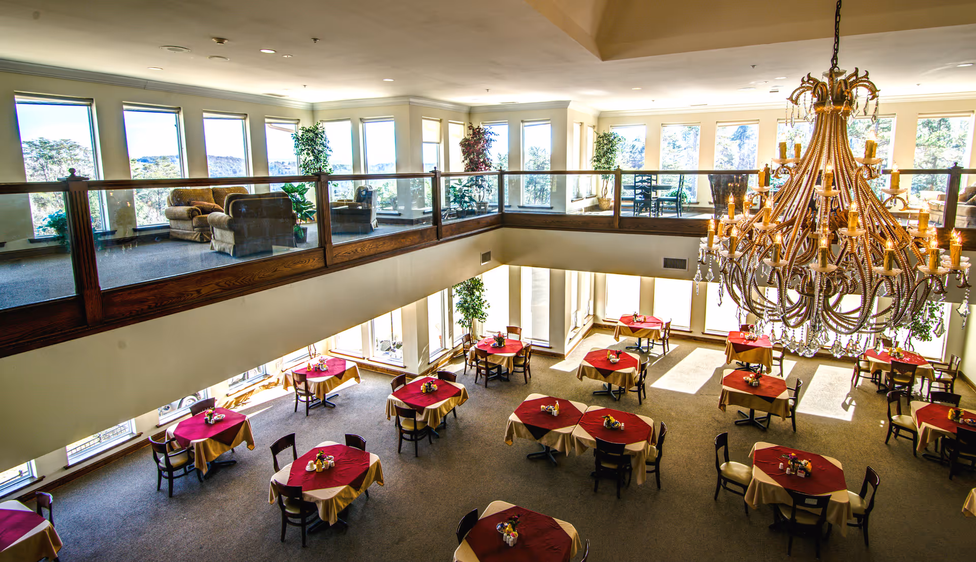 Spacious dining room with round tables draped in red tablecloths, a large chandelier, and an upper-level lounge with sofas.