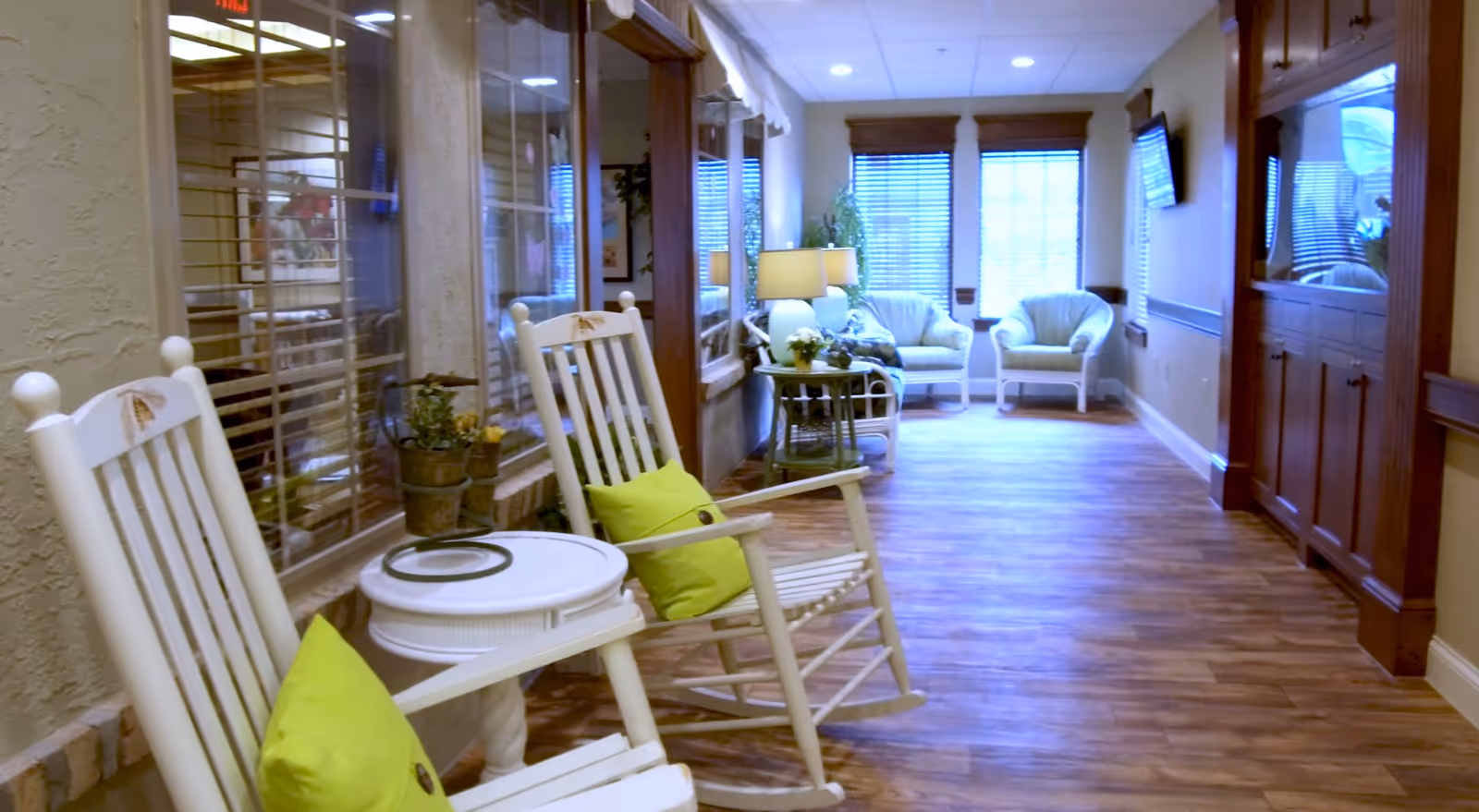 A bright sitting area in a senior living facility with white rocking chairs and a small table leading to armchairs and a wall-mounted TV.