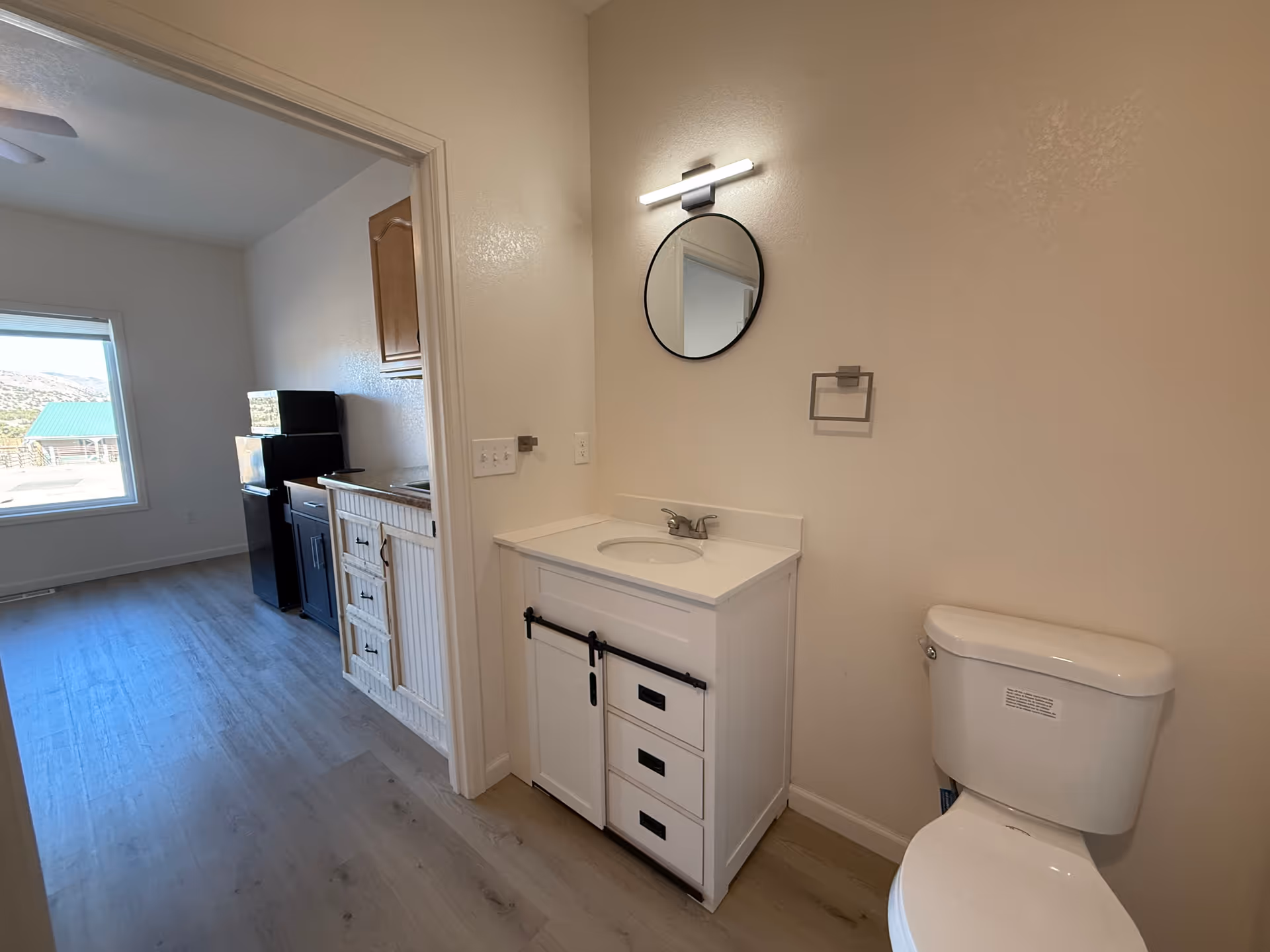 A small bathroom area with a white toilet and a white vanity with a sink. Above the vanity is a round mirror with a horizontal light fixture. To the left, there is an open doorway leading to a room with a window, a black refrigerator, a microwave, and wooden cabinets. The floor is light-colored wood, and the walls are painted white.