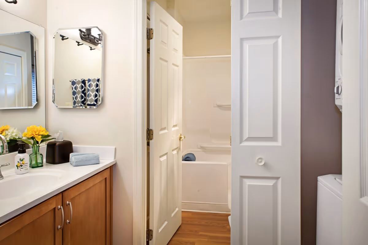 Bathroom with a wooden vanity and sink, mirrors and flowers, with a bathtub visible through an open door.
