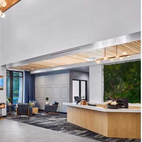 Modern senior living facility lobby with a wooden reception desk, a green moss wall art behind the desk, comfortable seating area with armchairs and small tables, large windows with blue curtains, and a high ceiling with wooden paneling and contemporary lighting fixtures.