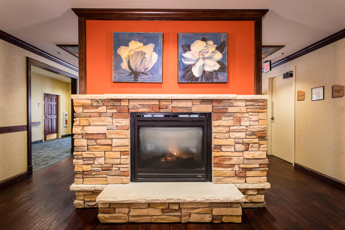 A cozy indoor stone fireplace with a small fire burning inside. Above the fireplace, there are two framed paintings of white flowers on a red-orange wall. The surrounding area has wooden flooring and beige walls with dark wood trim. There are doorways and hallways visible on either side of the fireplace.