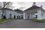Exterior view of a single-story building with gray siding and white trim, featuring a covered entrance with white columns and a cupola on top. The driveway curves around the entrance, and there are trees and grass surrounding the building under a cloudy sky.