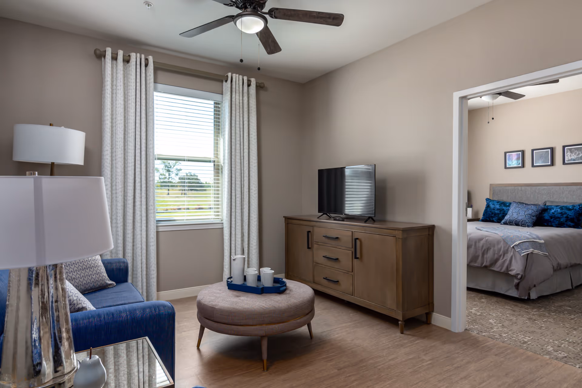 A cozy living room with a blue armchair, a round ottoman with a tray holding cups and a pitcher, a wooden TV stand with a flat-screen television, and a window with white curtains. The room has beige walls and wood flooring. Through an open doorway, a bedroom with a bed, blue pillows, and framed pictures on the wall is visible.