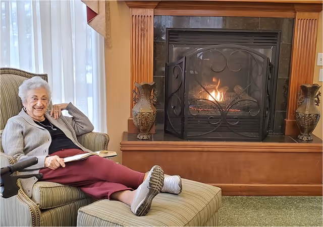 An elderly woman with white hair sits comfortably in a striped armchair with an ottoman, smiling and holding a book. She is wearing a gray cardigan, black top, and maroon pants. Behind her is a lit fireplace with a decorative black metal screen and two ornate vases on the mantel.