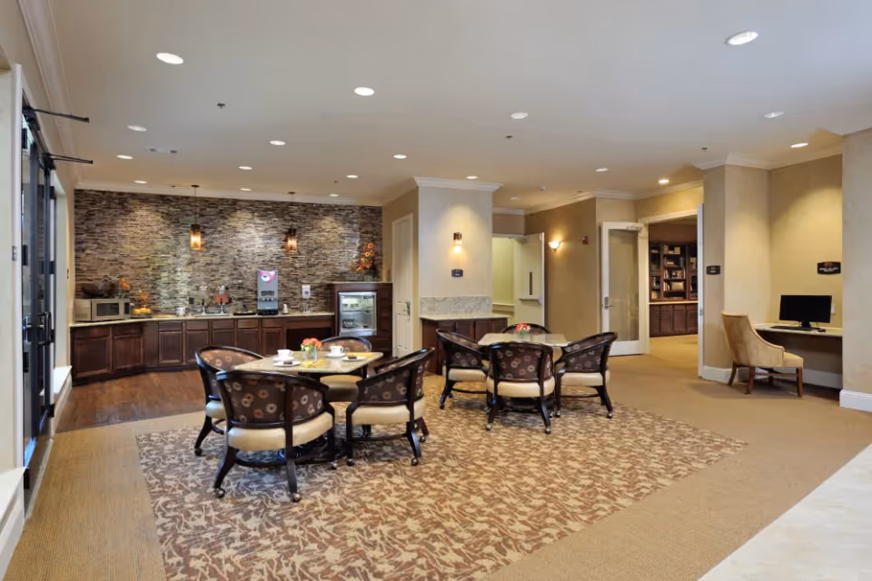 A cozy common area with two round tables surrounded by cushioned chairs on a patterned carpet. The back wall features a stone accent with a counter below holding a microwave, coffee machine, and a small refrigerator. To the right, there is a small desk with a computer and a chair. The room has warm lighting and beige walls.