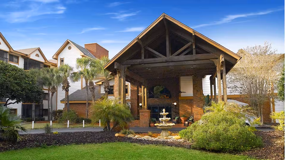 Exterior view of Vitality Living Baypoint Village showing a covered entrance with wooden beams and brick pillars, surrounded by palm trees, shrubs, and a small fountain in the foreground under a clear blue sky.