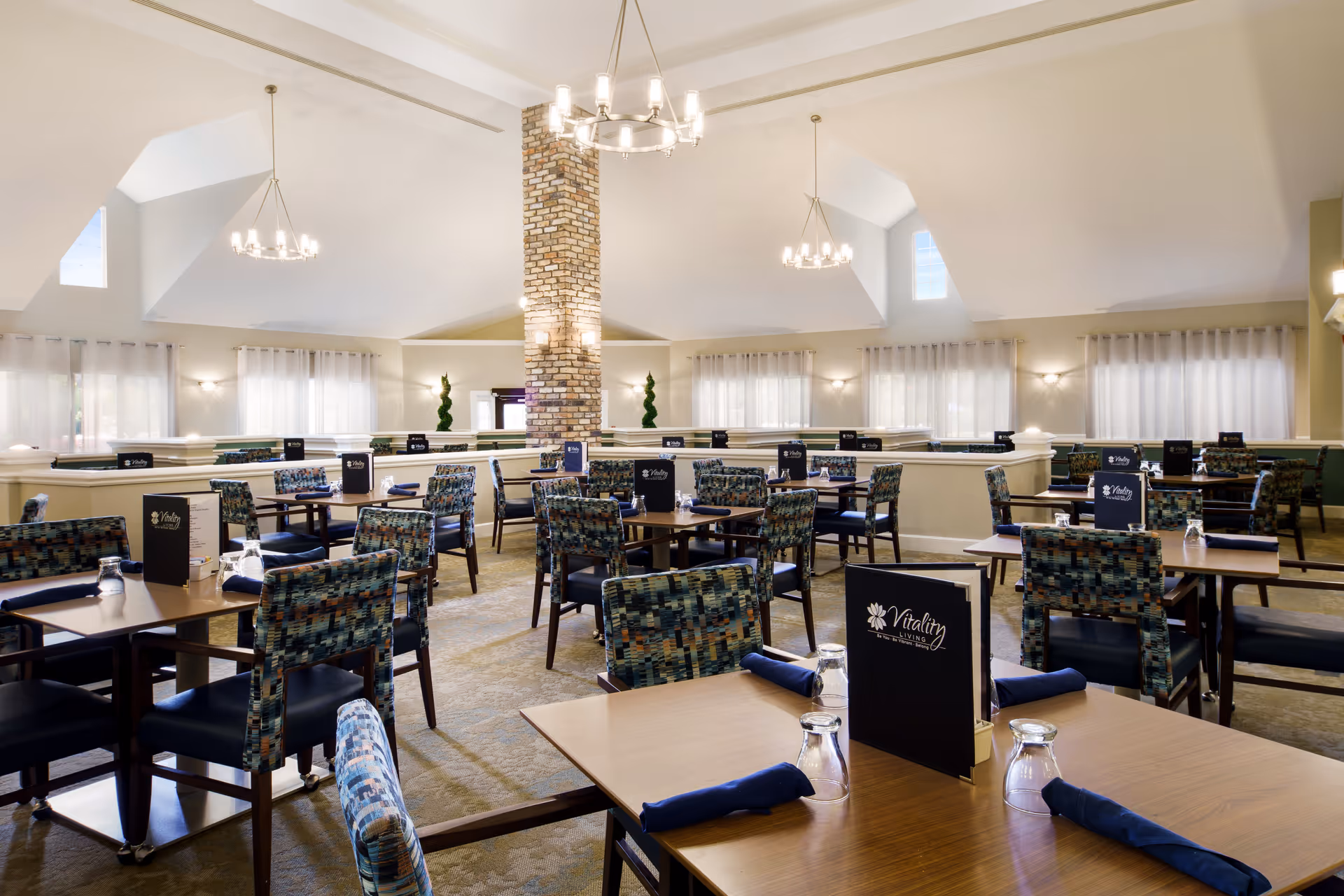 A spacious dining room with multiple wooden tables and patterned chairs arranged neatly. Each table has a menu labeled 'Vitality Living' along with blue napkins and upside-down glasses. The room features high vaulted ceilings with modern chandeliers, large windows with sheer curtains, and a central brick column.