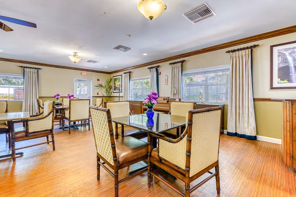A bright and spacious dining room with multiple tables and cushioned chairs. Each table has a small vase with purple flowers. The room features large windows with cream-colored curtains, wooden flooring, and light yellow walls with wooden trim. A piano and framed artwork are visible in the background.