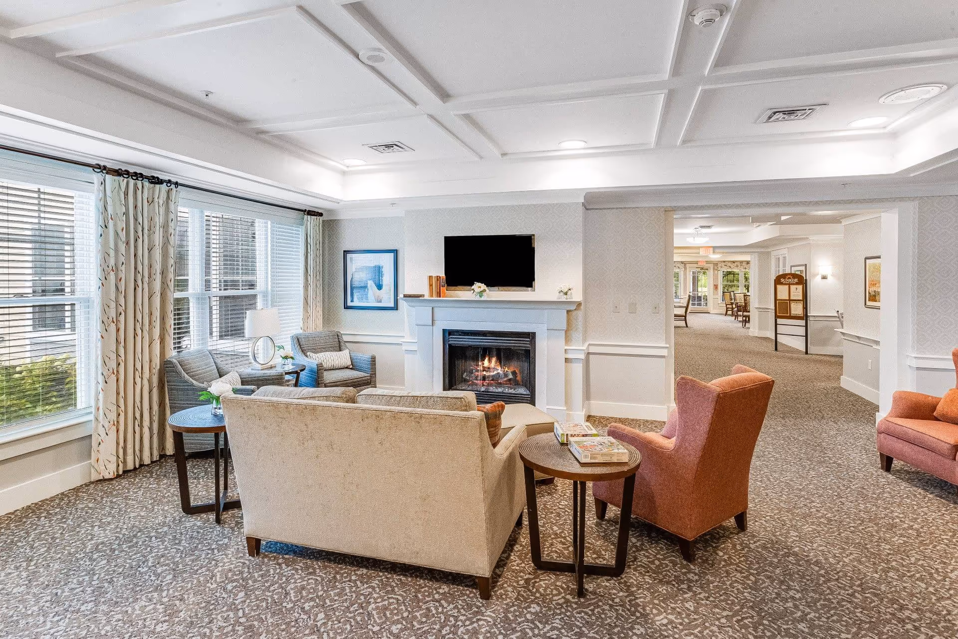 A cozy living room area in a senior living facility with a beige sofa, two orange armchairs, and two gray armchairs arranged around a fireplace with a mounted flat-screen TV above it. There are large windows with patterned curtains letting in natural light, a side table with a lamp, and a carpeted floor with a patterned design. The room opens into a hallway with more seating and decor visible in the background.