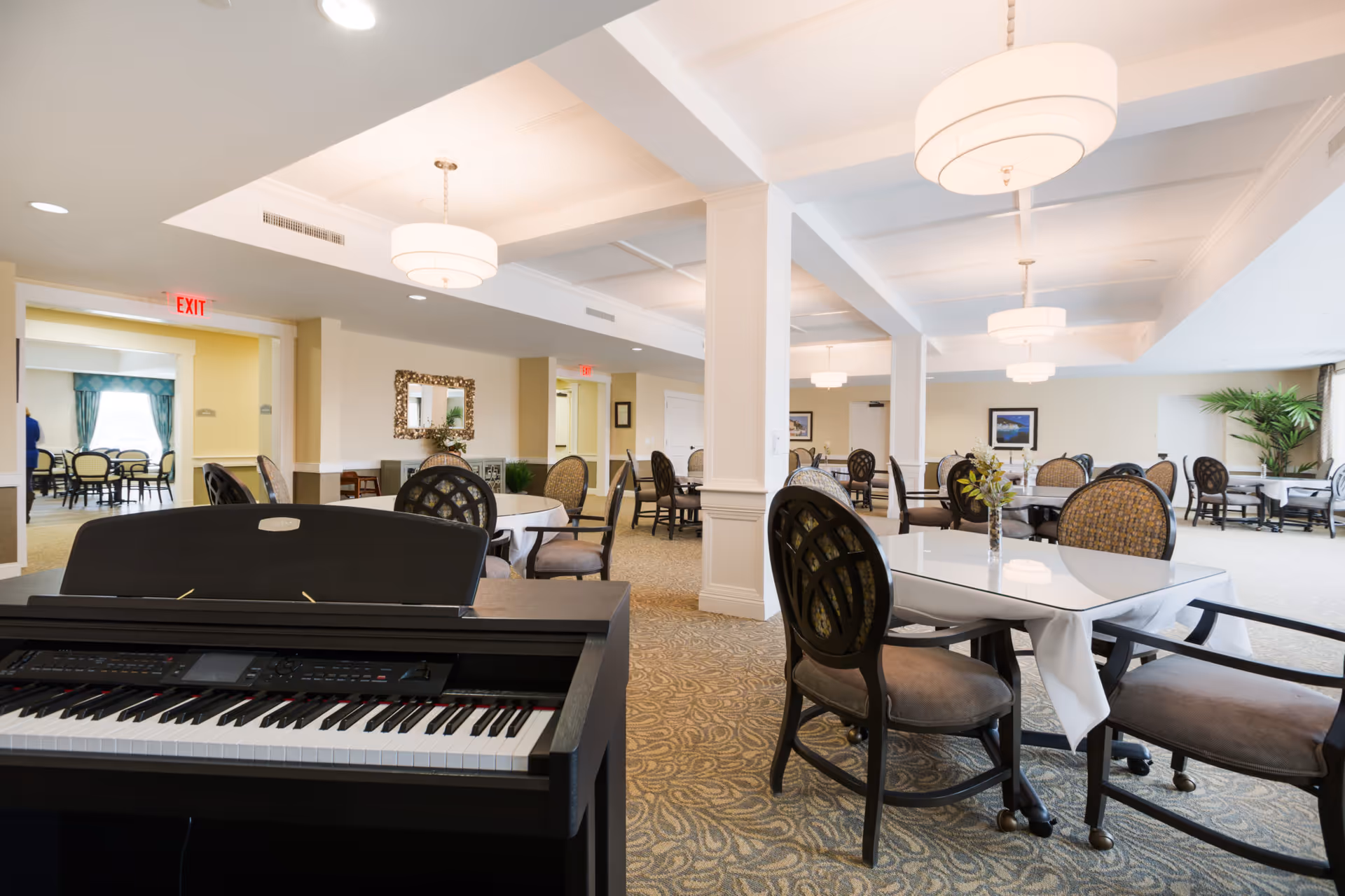 A spacious dining room in a senior living facility with multiple tables covered in white tablecloths and surrounded by cushioned chairs. A black electronic keyboard is positioned in the foreground. The room is well-lit with modern ceiling lights and has beige walls and patterned carpet. There are plants and framed pictures on the walls, and an exit sign is visible in the background.