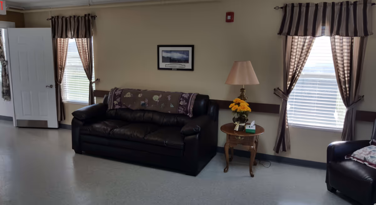 A living room area with a black leather couch and a matching armchair. The couch has a decorative throw blanket draped over the back. Between the couch and armchair is a small wooden side table with a lamp, a vase of yellow flowers, and a box of tissues. Two windows with brown striped curtains allow natural light into the room. A framed picture hangs on the beige wall above the couch. The door to the left is open, revealing part of the hallway.