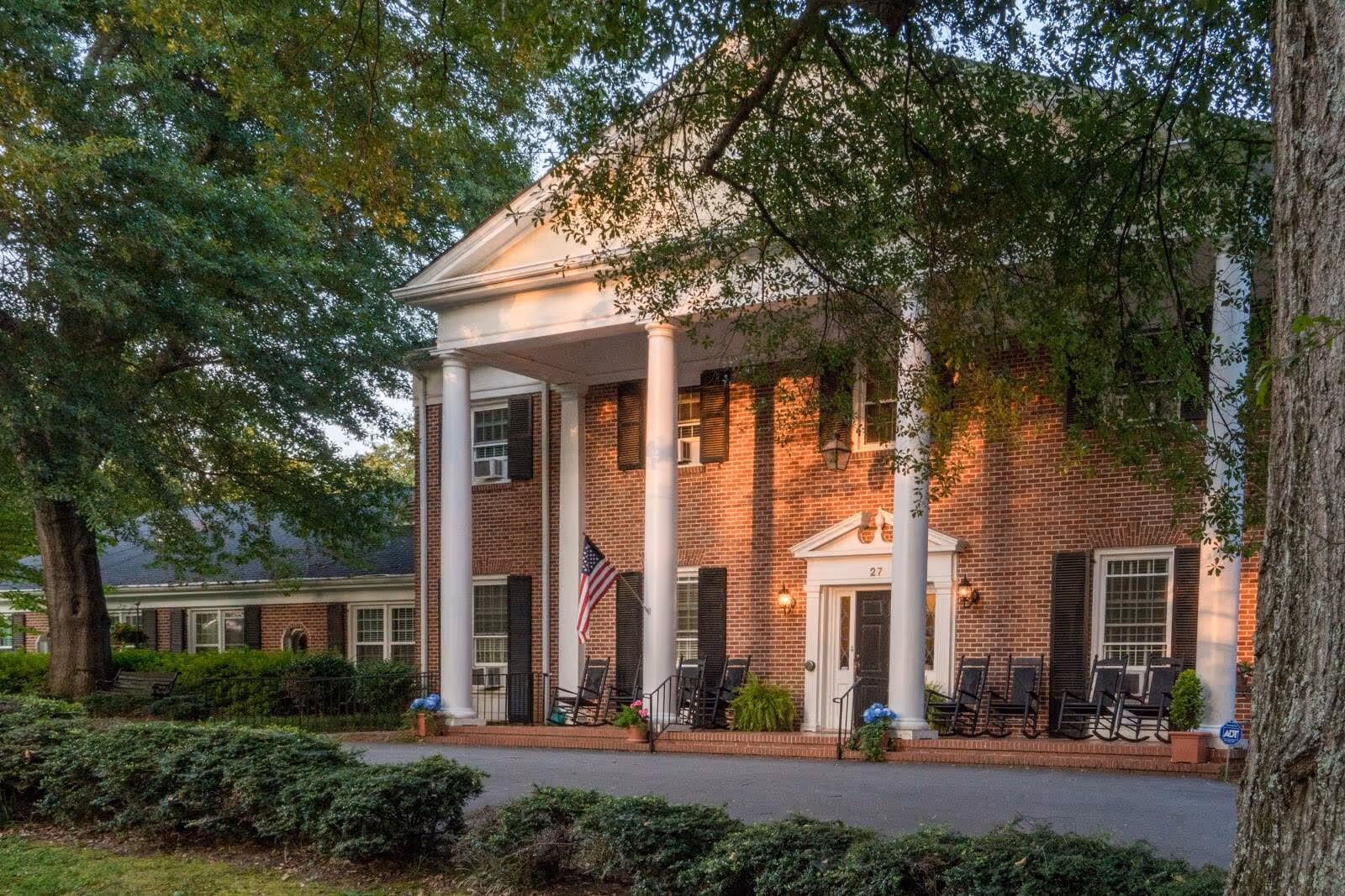 Front exterior view of a two-story brick building with white columns and a covered porch. Several black rocking chairs are arranged on the porch, and an American flag is displayed near the entrance. The building is surrounded by trees and greenery.