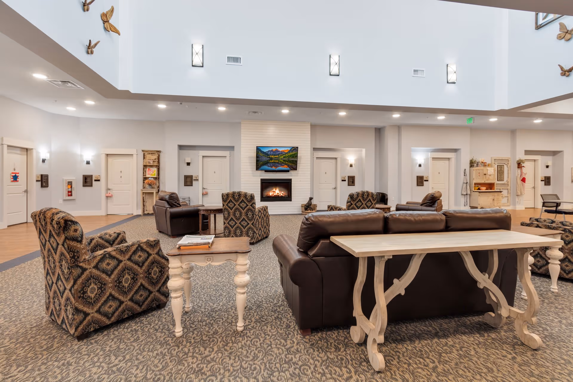 Spacious senior living facility common area with patterned armchairs, leather sofas, a wooden table, and a fireplace with a TV mounted above it. The room has high ceilings with wall sconces and several doors leading to other rooms. Decorative butterflies are mounted on the walls.