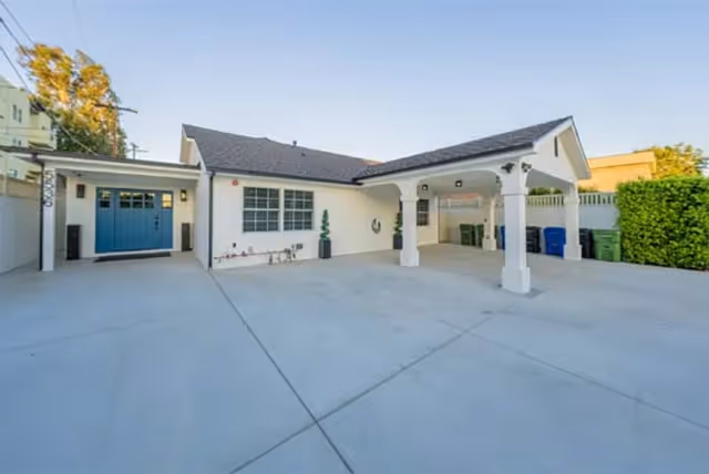 Exterior view of a single-story building with a covered carport supported by white columns, a blue garage door, and a large concrete driveway. There are some green trash bins lined up against a fence on the right side and a hedge on the far right.