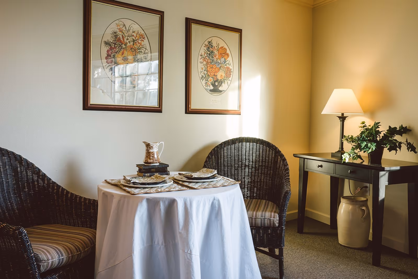 A cozy dining area with a small round table covered with a white tablecloth, set for two with plates, napkins, and a decorative pitcher on top of stacked books. Two wicker chairs with striped cushions are placed on either side of the table. On the right, there is a black console table with a lamp and a potted plant, and a large ceramic jar underneath. Two framed floral prints hang on the wall above the table.