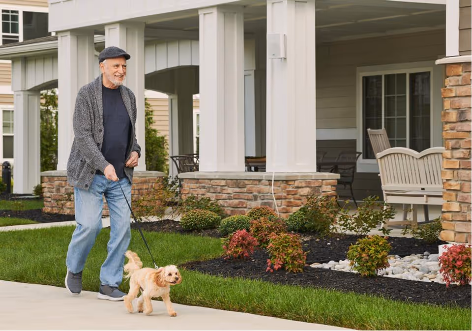 An elderly man wearing a gray cardigan, blue jeans, and a black cap is walking a small light brown dog on a leash outside a senior living facility. The background shows a porch area with white columns, brick accents, outdoor chairs, and landscaped bushes.