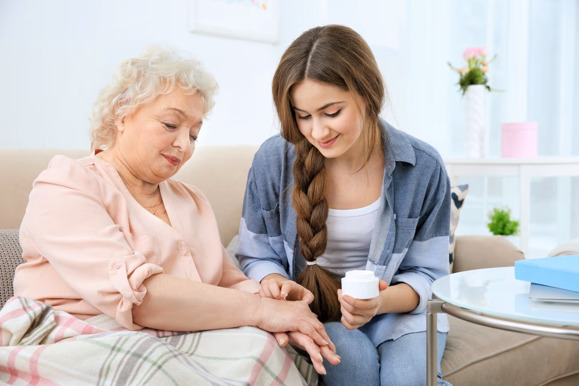 A young woman with a long braid is sitting on a couch next to an elderly woman with short curly hair. The young woman is holding a jar of cream and gently applying it to the elderly woman's hand. They are in a bright, cozy living room with a blanket draped over the elderly woman's lap and a round glass table nearby with some books and a plant in the background.