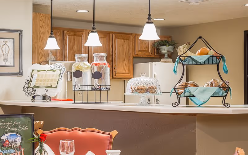 Interior view of a kitchen and dining area in a senior living facility. The kitchen has wooden cabinets, a white refrigerator, and a countertop with two beverage dispensers filled with lemon and berry water. There is a glass cake stand with muffins and a two-tiered metal stand holding fruit and pastries. Three pendant lights hang above the counter. In the foreground, part of a dining table with a red cushioned chair, a wine glass, and a menu is visible.