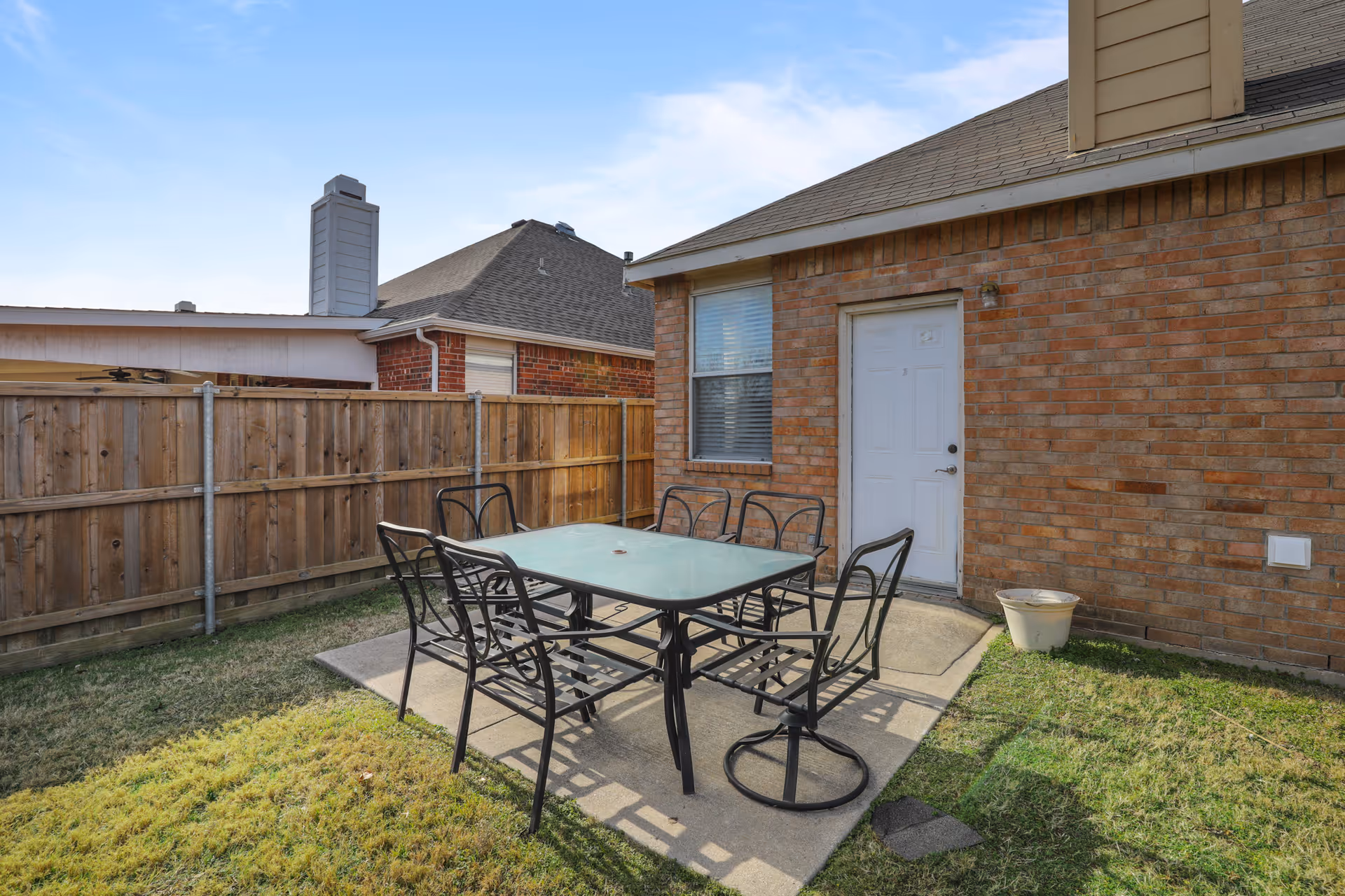 Outdoor patio area with a glass-top table and six black metal chairs on a concrete slab, surrounded by grass and a wooden fence. A brick building with a white door and window is visible in the background under a partly cloudy sky.