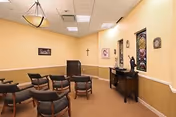 A small meeting or chapel room with beige walls and carpeted floor. The room contains several wooden chairs with cushions arranged in rows facing a wooden podium. On the right wall, there is a console table with decorative items and a stained glass window. A cross and framed pictures are also visible on the walls.