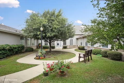Outdoor courtyard area at The Wellington at Arapaho featuring a large tree in the center surrounded by a curved concrete walkway, potted plants with flowers, two wooden chairs on the grass, and single-story building walls with windows in the background under a blue sky with some clouds.