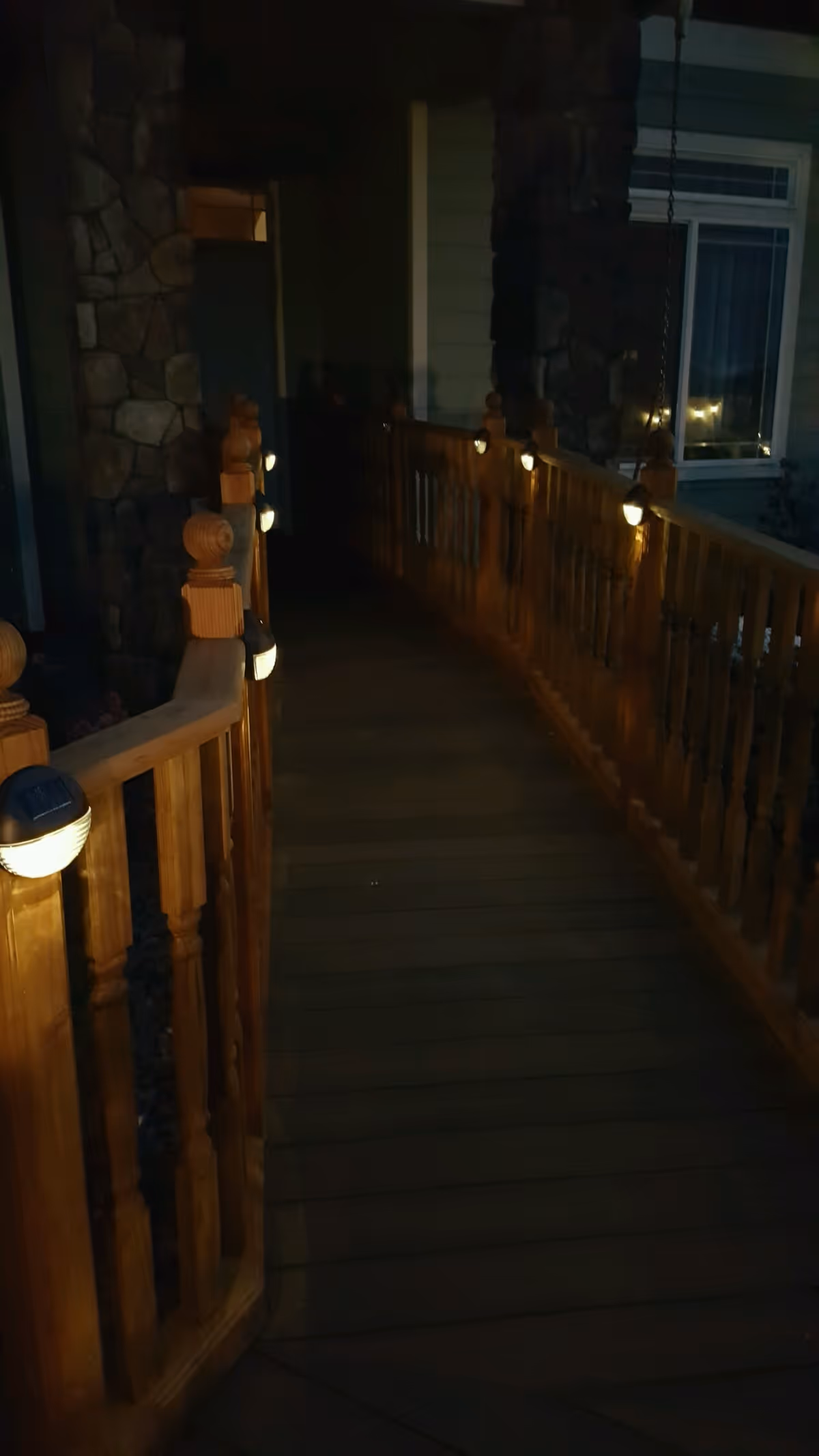 A wooden ramp with railings on both sides, illuminated by small lights attached to the railings, leading to the entrance of a building at night. The building exterior features stone and siding with a window visible on the right side.