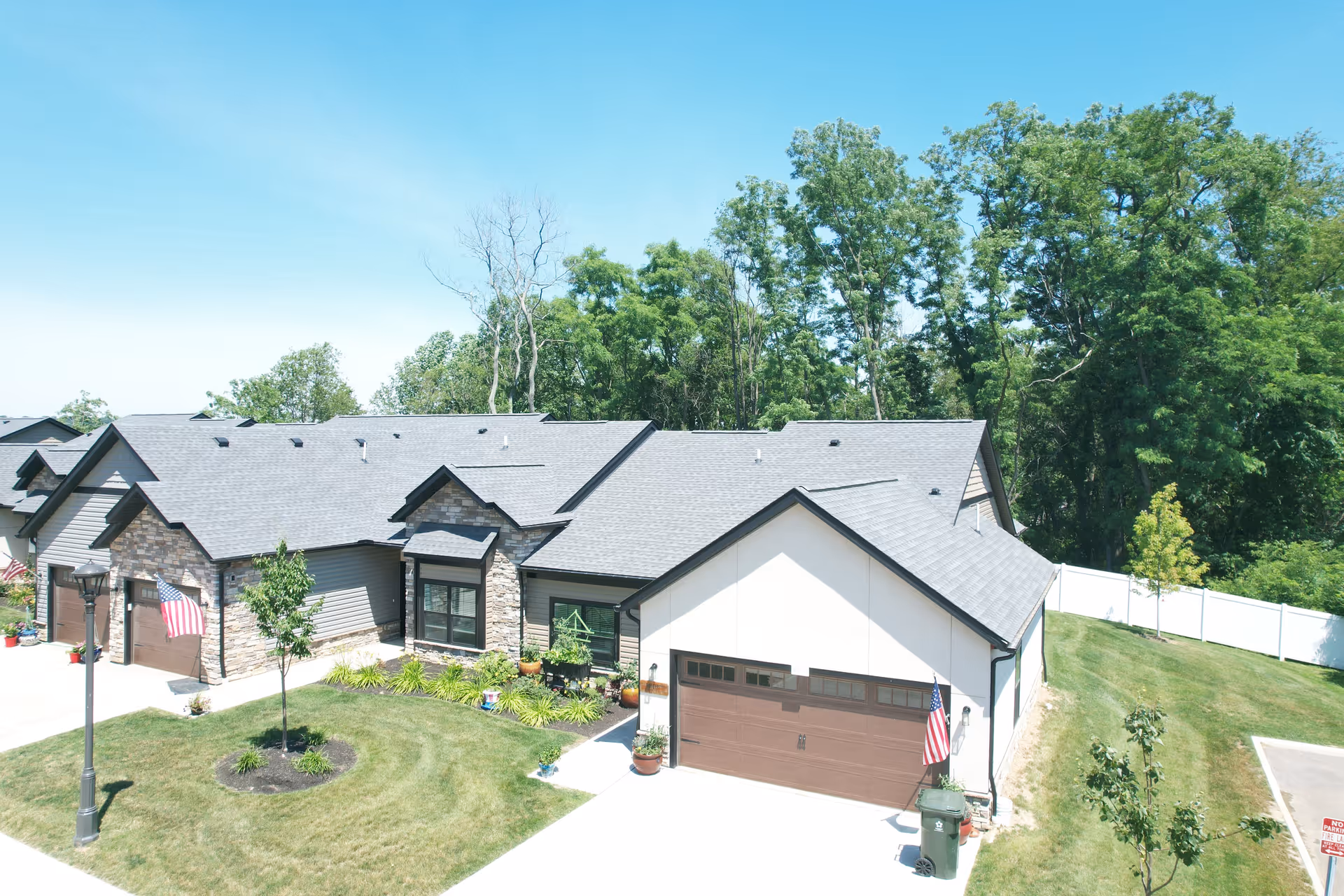 Exterior view of a single-story residential building with a gray roof, stone and siding facade, attached garage with a brown door, and American flags displayed near the garage and driveway. The building is surrounded by a well-maintained lawn, small trees, and a white fence with tall green trees in the background under a clear blue sky.