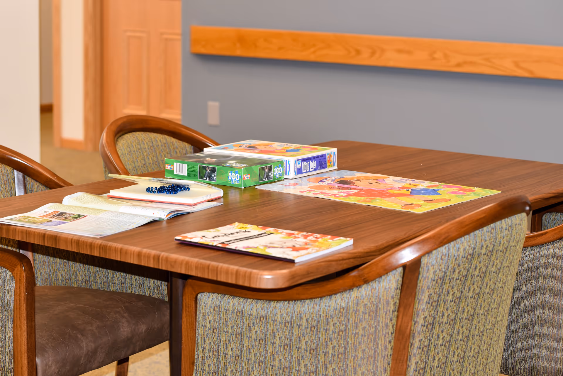 A wooden table with four upholstered chairs around it in a room with light blue walls and a wooden trim. On the table are two jigsaw puzzles, an open magazine, a notebook with a beaded bracelet on top, and a colorful placemat featuring a cartoon character.