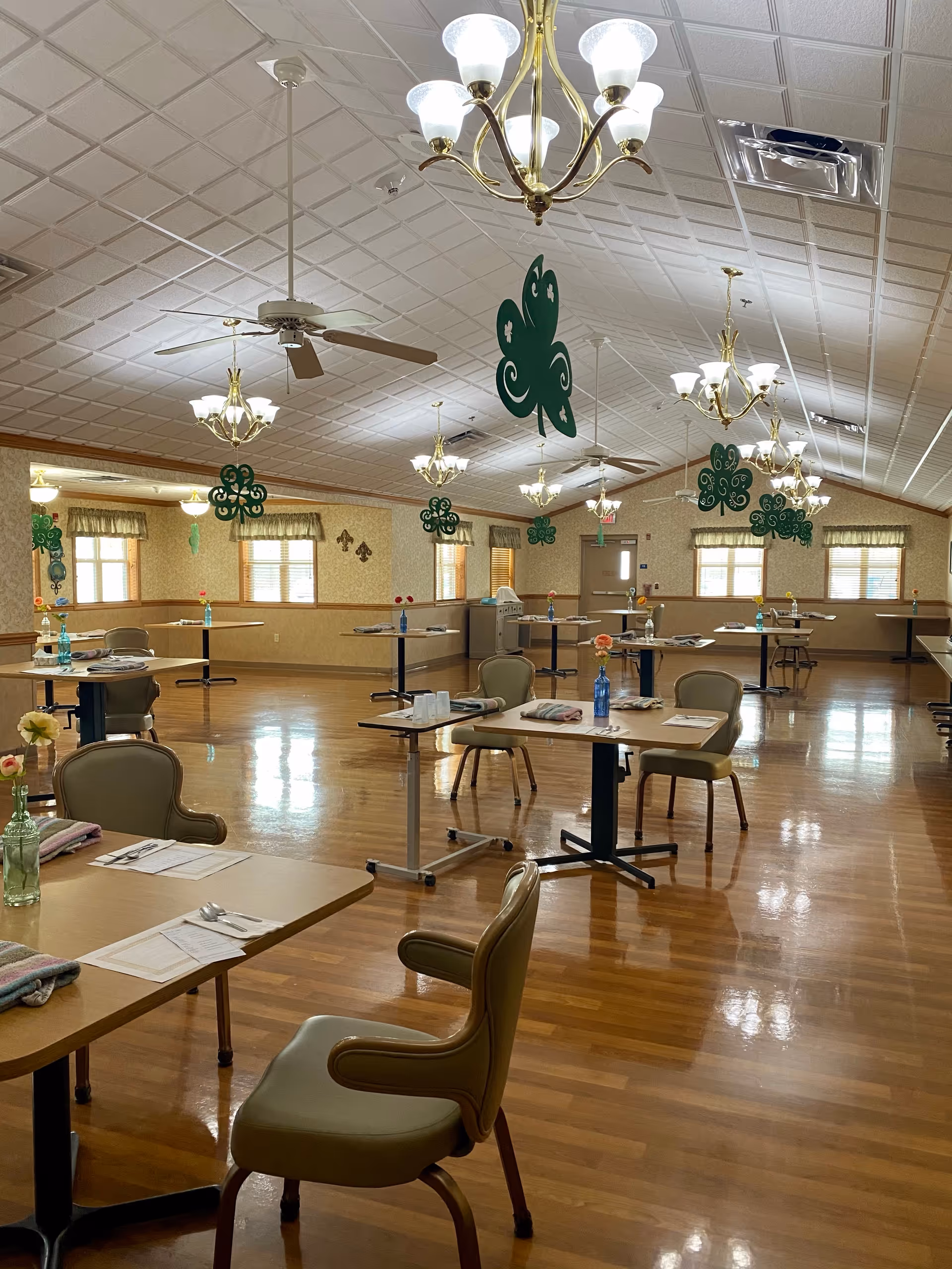A spacious dining room with wooden floors and several tables set with placemats, utensils, and single flowers in vases. The ceiling has multiple chandeliers and ceiling fans, and green shamrock decorations hang from the ceiling. Windows with curtains line the walls, and the room is well-lit with natural and artificial light.